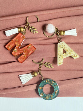 A collection of charms with the letters M, A, and O are arranged on a pink background. The charms are made of resin and have a gold finish, giving them a luxurious and elegant appearance