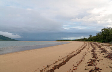A quiet curve of untouched beach meets still blue water under a moody, overcast sky.