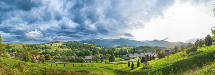 Scenic view of a Carpathian village surrounded by lush green hills, haystacks, forests, and dramatic clouds. Traditional rural landscape in the Ukrainian mountains.