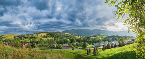 Scenic view of a Carpathian village surrounded by lush green hills, haystacks, forests, and dramatic clouds. Traditional rural landscape in the Ukrainian mountains.