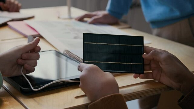 Close-up of hands of diverse young students examining small scaled solar battery and plug for powering digital tablet computer, while learning about green energy sources from teacher