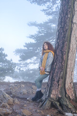 A young female traveler standing in a misty forest, surrounded by tall trees and dense fog. She wears a backpack and outdoor clothing, blending into the mysterious, atmospheric landscape. 