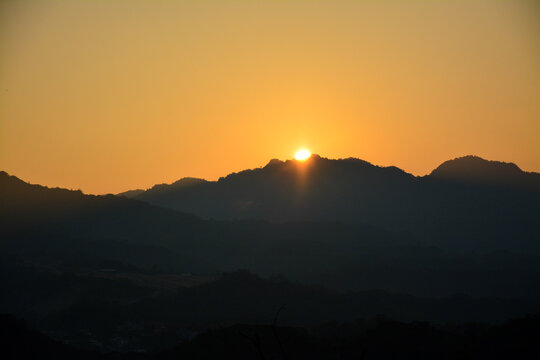 Sun Emerging Over Mountain Range at Dawn: A view of the sun just peeking over the horizon of a silhouetted mountain range against a warm-toned morning sky.