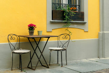 Chairs and a table are set against the yellow wall at an outdoor cafe in Alba, Italy.