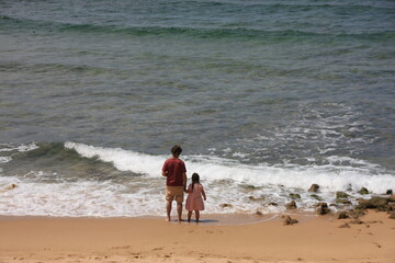 Father stands on the beach with his young daughter and looks out to sea