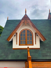 Close-up of a dormer window with a green shingle roof, white trim, and ornate wooden details. Cloudy sky above.