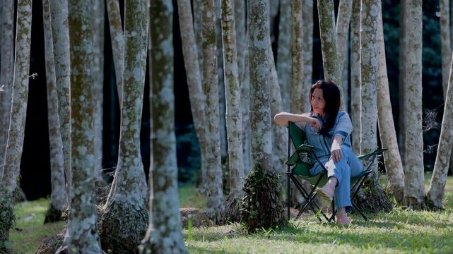 Confident smiling middle aged asian woman, 49 years old lady relaxing on a camping chair admiring the magnificent landscape of the forest. The sun beautifully illuminating the tree. 