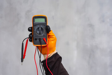 Electrician wearing a protective glove, holding a digital multimeter. Orange colored instrument with connected test leads for measuring voltage, current and resistance in electrical systems.