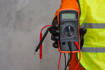 Electrician wearing a protective glove, holding a digital multimeter. Orange colored instrument with connected test leads for measuring voltage, current and resistance in electrical systems.