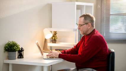 An elderly man is sitting at his desk working with a laptop at home.