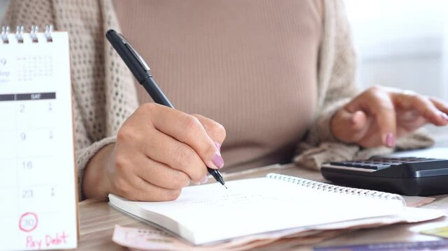 Close-up of a woman's hand writing in a notebook and using a calculator to manage her monthly expenses and debt payments, showing personal budgeting and financial planning at home
