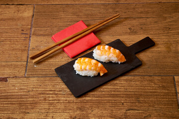 Two pieces of sushi on a black tray. The tray is on a wooden table. There are chopsticks next to...