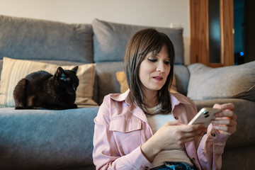 Woman using smartphone at home with black cat resting on sofa