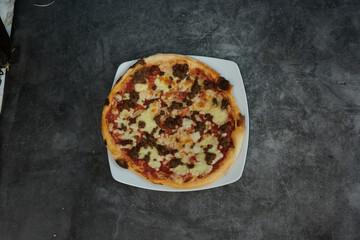 A pizza with cheese and meat toppings sits on a white plate. The pizza is cut into four pieces