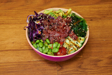 A bowl of food with a variety of vegetables and a piece of meat. The bowl is pink and is placed on a wooden table