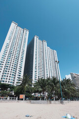 Modern high-rise apartment buildings near the beachfront in Nha Trang, Vietnam. The photo captures a clear blue sky, palm trees, and part of the sandy beach in the foreground.

