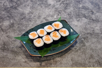 A plate of sushi with a green leaf on top. The sushi is cut into six pieces. The plate is on a grey surface