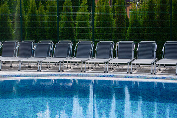 Empty deck with swimming pool blue water in summer. Pool side view against the background of green bushes. Sun loungers. Start of the season