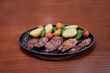 A plate of meat and vegetables on a wooden table. The meat is cut into small pieces and is served on a black plate. The vegetables include potatoes, tomatoes, and peppers