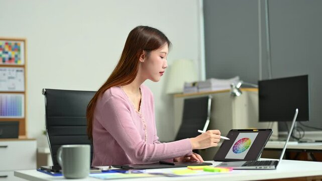 Young female designer using digital tablet and laptop in a modern creative office. Surrounded by colors, notes, and ideas.