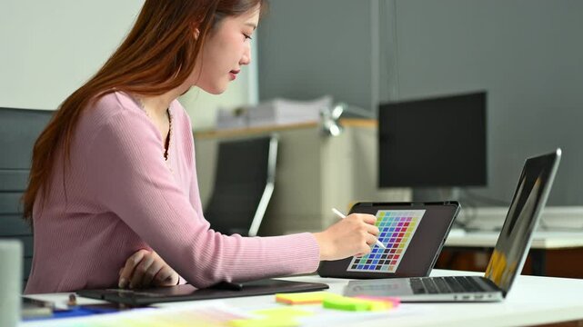 Young graphic designer using pen tablet and laptop at creative desk in modern office. Surrounded by color palettes and design tools.