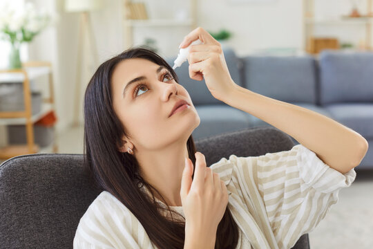 Focused young woman leaning back in armchair, holding dropper and applying eye drops. Female treating dry, irritated or inflamed eyes, focusing on health care routine and maintaining eye moisture.