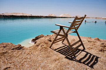 Wooden deck chair stands near water edge of salt lake. Unrecognizable people swim in lake at background. Turquoise water and white salt on the shore of Siwa salt lake, Western Desert, Egypt