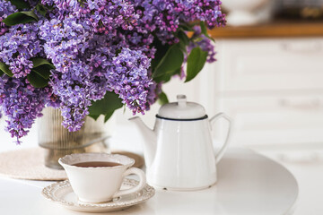 A large purple bouquet of lilacs, white Scandinavian houses, a ceramic teapot and a cup on the table. In the background is the interior of a modern white kitchen. The concept of home comfort.
