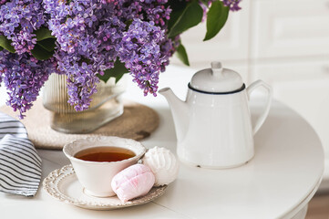 A large purple bouquet of lilacs, a ceramic teapot, a cup and a delicious marshmallow on the table. In the background is the interior of a modern white kitchen. The concept of home comfort.