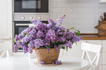 A large purple bouquet of lilacs on the table in a wicker basket. In the background is the interior of a modern white kitchen. The concept of home comfort and celebration.
