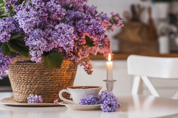 A large purple bouquet of lilacs in a wicker basket, a cup of tea and candles on the table. In the background is the interior of a modern white kitchen. The concept of home comfort.