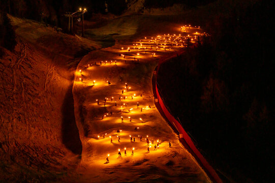 A high-angle shot shows a torchlight procession snaking down a snowy slope at night. Many small, bright lights illuminate the path. Monte Santo di Lussari, Tarvisio, Friuli Venezia Giulia, Italy.