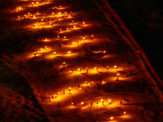 A high-angle shot shows a torchlight procession snaking down a snowy slope at night. Many small, bright lights illuminate the path. Monte Santo di Lussari, Tarvisio, Friuli Venezia Giulia, Italy.