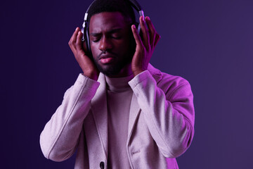 A young Black man enjoying music with headphones on, dressed in a stylish beige coat, against a...