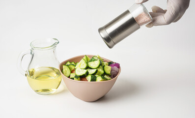Prepared vegetable salad in a bowl on a white background. Nearby is olive oil in a glass jug. A woman's hand adds salt to the salad