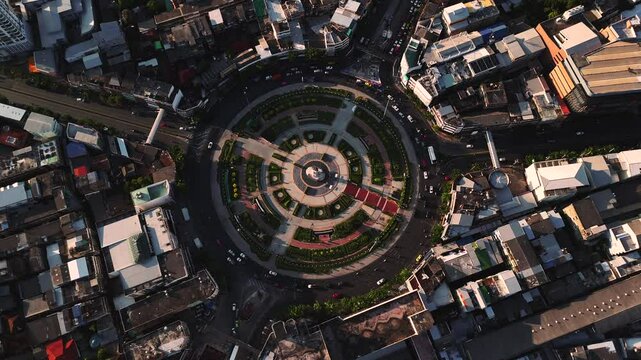 Drone top-down view of Wongwian Yai traffic circle in Thon Buri district, Bangkok. The iconic roundabout with King Taksin monument is surrounded by low-rise urban buildings at sunset