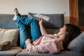 Young woman lying on sofa playing with paper airplane: dreaming of future travel and new experiences