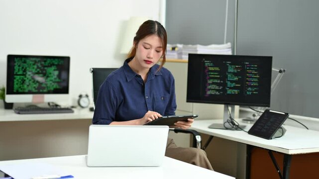 Young female programmer working with tablet and screen displaying code. Modern tech office setup, great for software, coding, IT