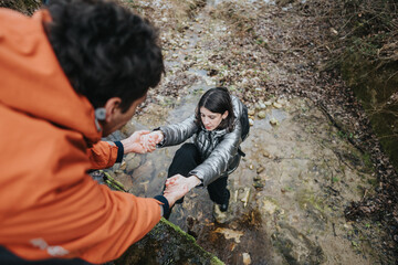 Individuals hiking in an outdoor forest environment demonstrate assistance and teamwork while navigating a rocky creek.