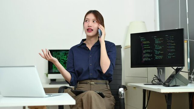 Young female developer talking on the phone while coding in a modern tech office. Surrounded by monitor, tablet, and laptop. Ideal for tech, IT