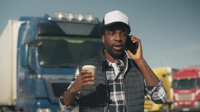 Serious African American male wearing cap and warm vest while talking on smartphone near parked trucks. Holding takeaway coffee while explaining situation or discussing schedule. Communicating.