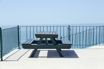 On a concrete terrace, a black wooden picnic bench faces the calm sea under a clear blue sky, sunny summer day. A metal railing frames the open view, emphasizing the peaceful and spacious atmosphere.