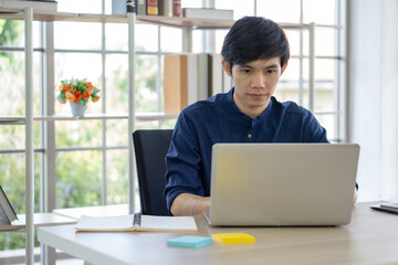 Fototapeta premium A focused young man type on his laptop while seated at a contemporary desk filled with notes and colorful sticky note, enjoying natural daylight streaming through large window.