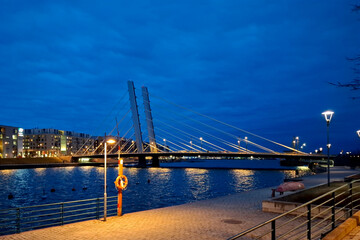 Illuminated modern cable‑stayed bridge spanning a calm river at dusk, with city lights reflecting on water and a riverside walkway under glowing street lamps.