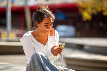 young woman sitting outdoors on a sunny day, laughing while looking at her phone