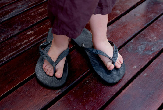Child's feet in oversized black flip-flops on dark wooden flooring.
