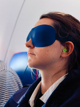 A woman with an eye mask and earplugs sits on an airplane seat, appearing relaxed.