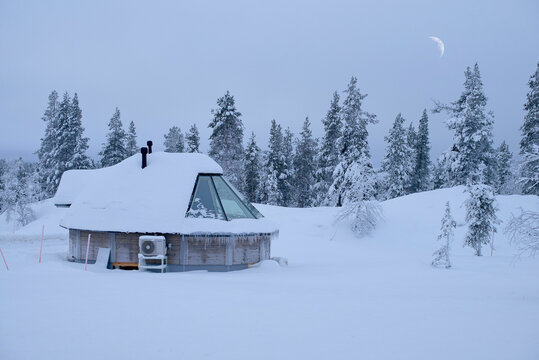Fototapeta Snow-covered wooden cabin with triangular window amidst snowy pine forest under crescent moon. North Finland