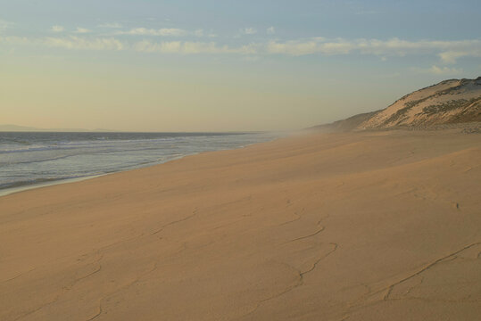 Sandy beach at sunset with gentle waves and a distant view of hills under a cloudy sky. Portugal