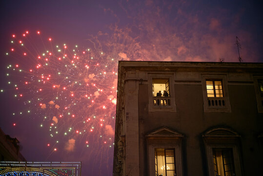 Fireworks light up the evening sky near an illuminated historic building with people watching. Sicily, Italy - Powered by Adobe